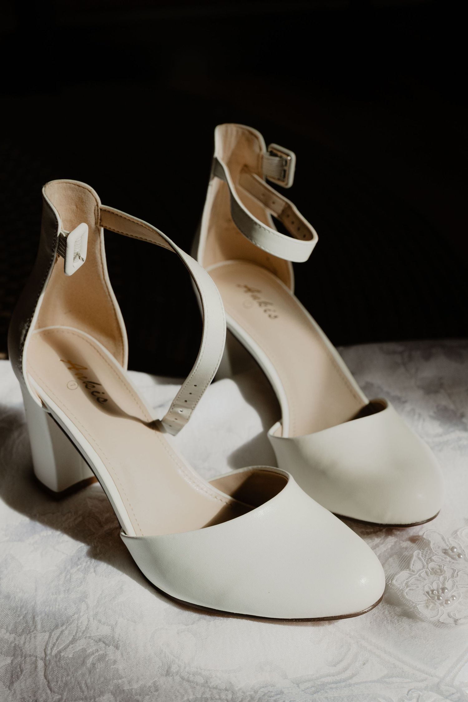 Elegant white block heel bridal shoes with ankle strap displayed on a lace surface.