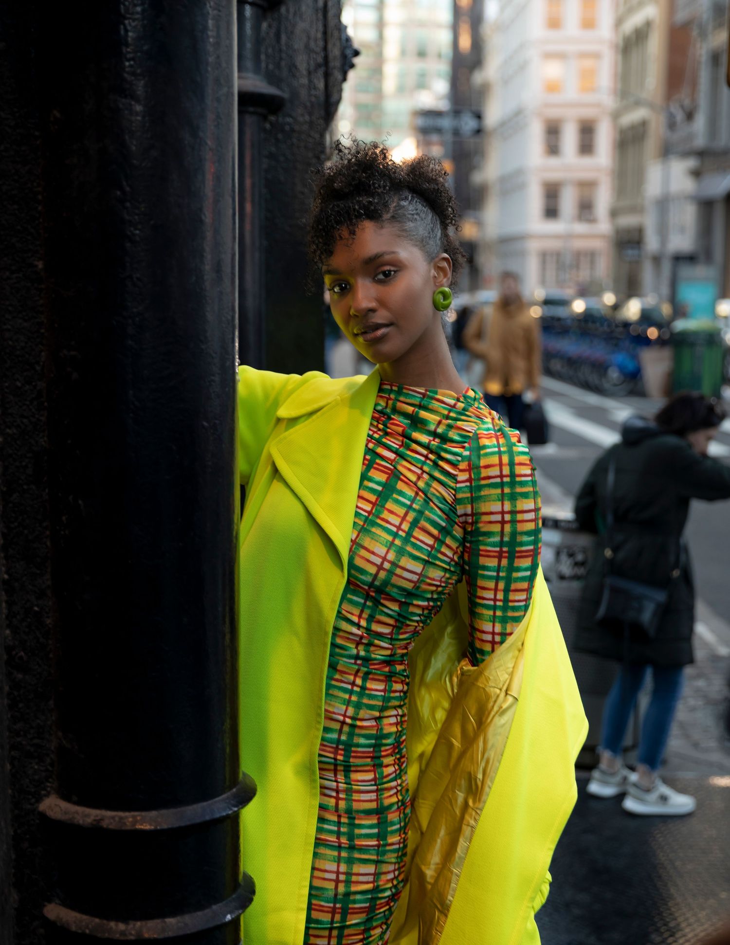 Woman in vibrant yellow coat and colorful kente print dress poses on urban street, showcasing bold fashion style.