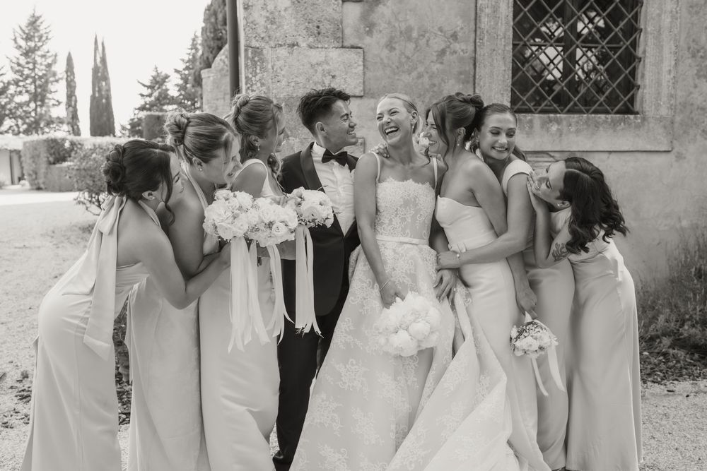 Black and white wedding photo of bride and groom laughing with bridesmaids in light gowns holding bouquets outdoors.
