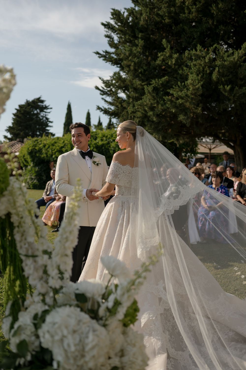 Bride in off-shoulder gown with long veil stands with groom in white tuxedo at outdoor wedding ceremony.
