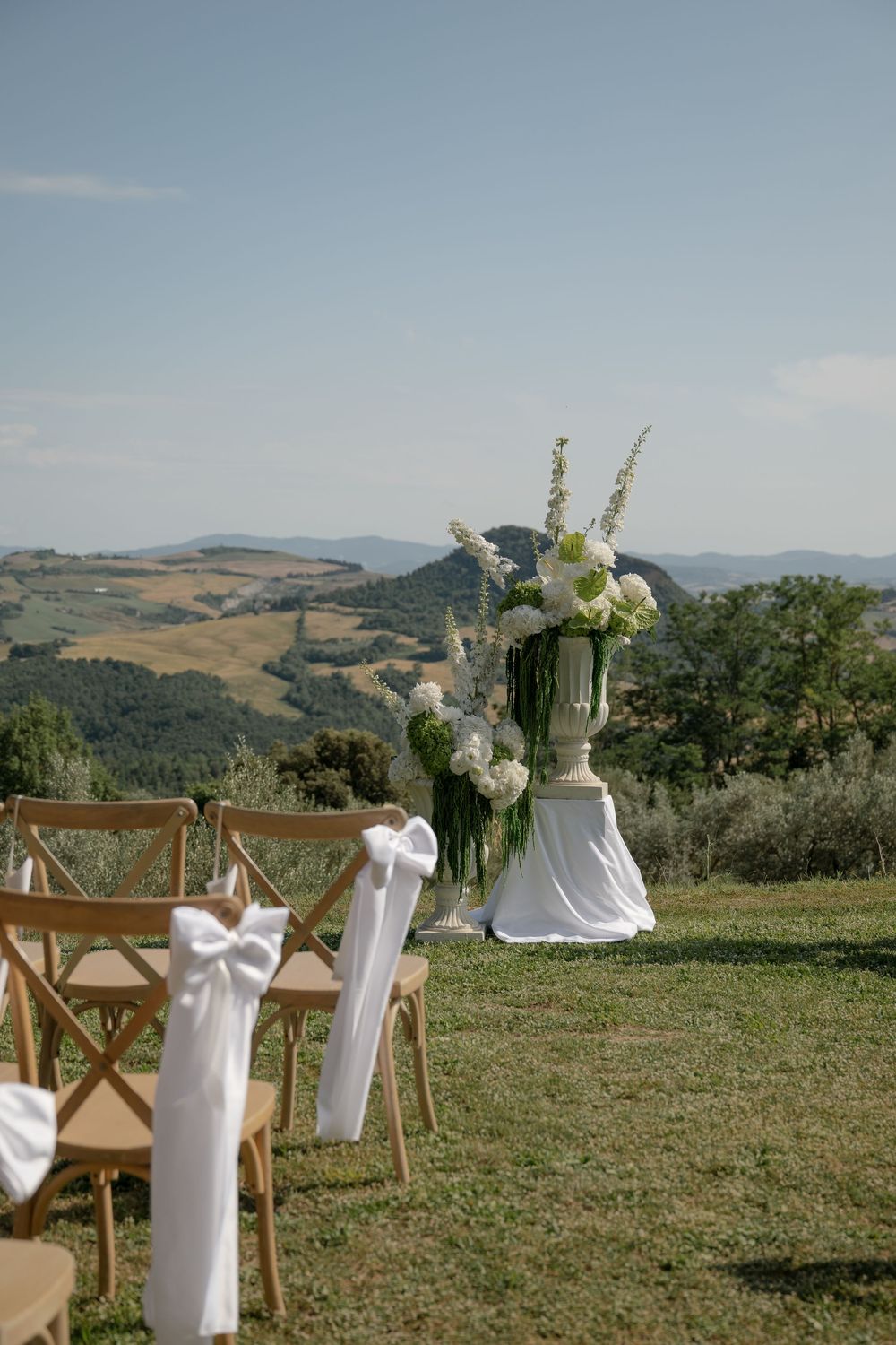 Outdoor wedding ceremony setup with wooden chairs, white floral urns, and scenic Tuscan hills backdrop.