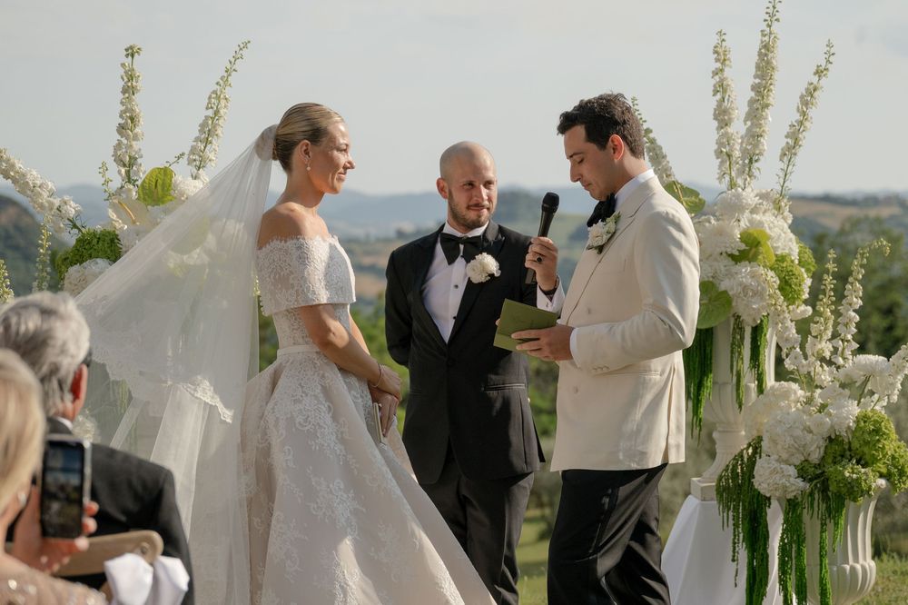 Elegant outdoor wedding ceremony with bride in lace gown, groom in white tuxedo, exchanging vows at floral arch.