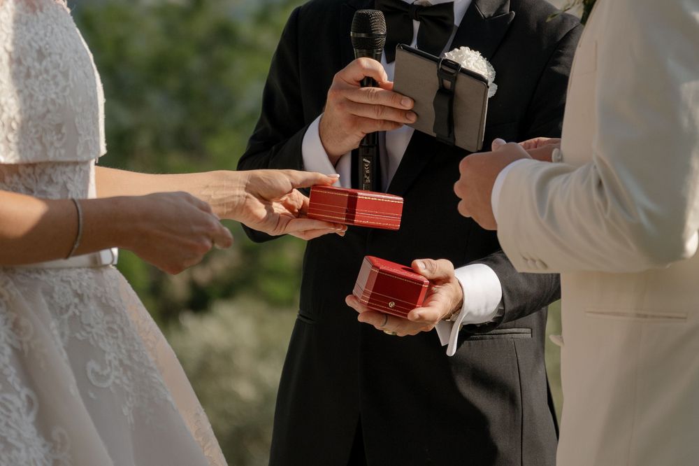 Bride and groom exchanging rings in red boxes during outdoor wedding ceremony with officiant holding microphone.