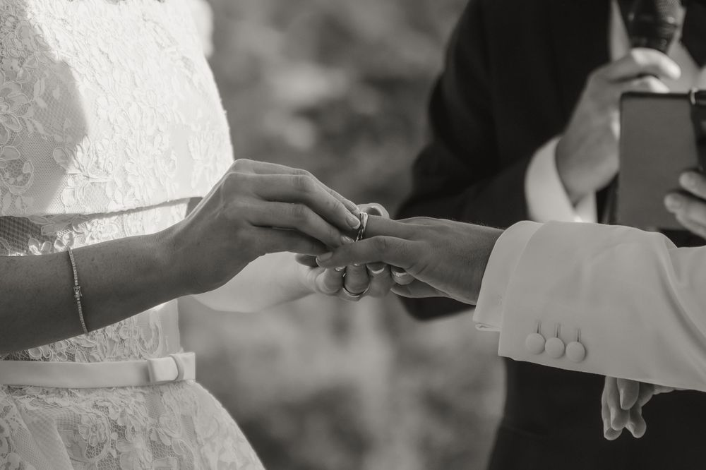 Black and white photo of bride placing wedding ring on groom's finger during outdoor ceremony.