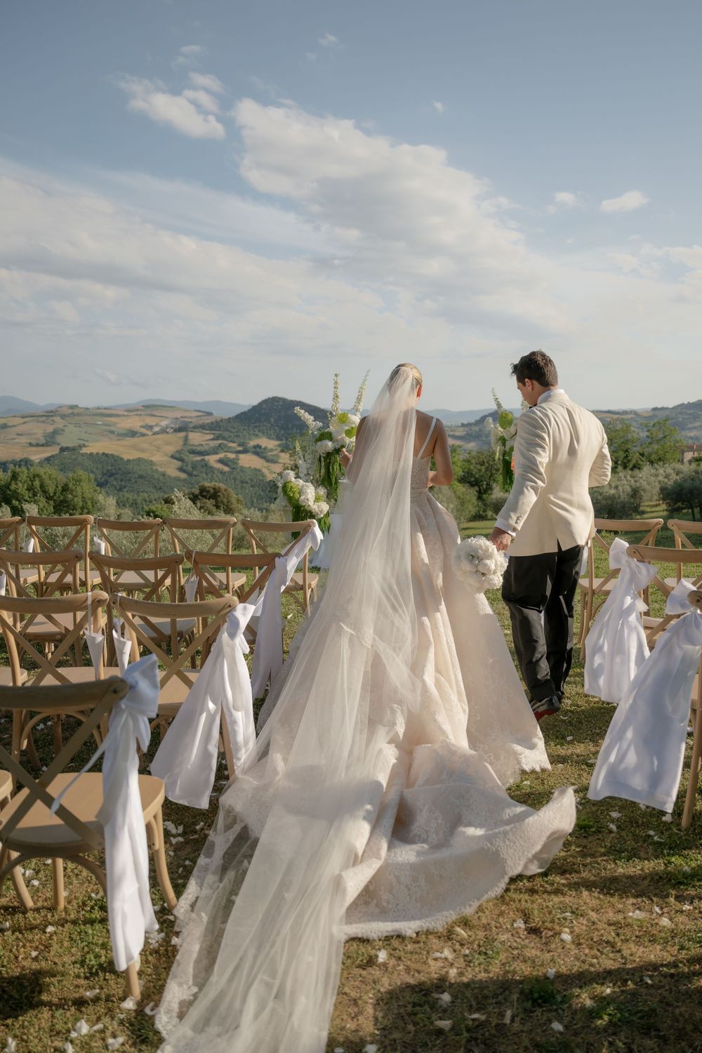 Bride and groom walk hand-in-hand at outdoor Tuscany wedding ceremony with rolling hills and white ribbon-adorned chairs.