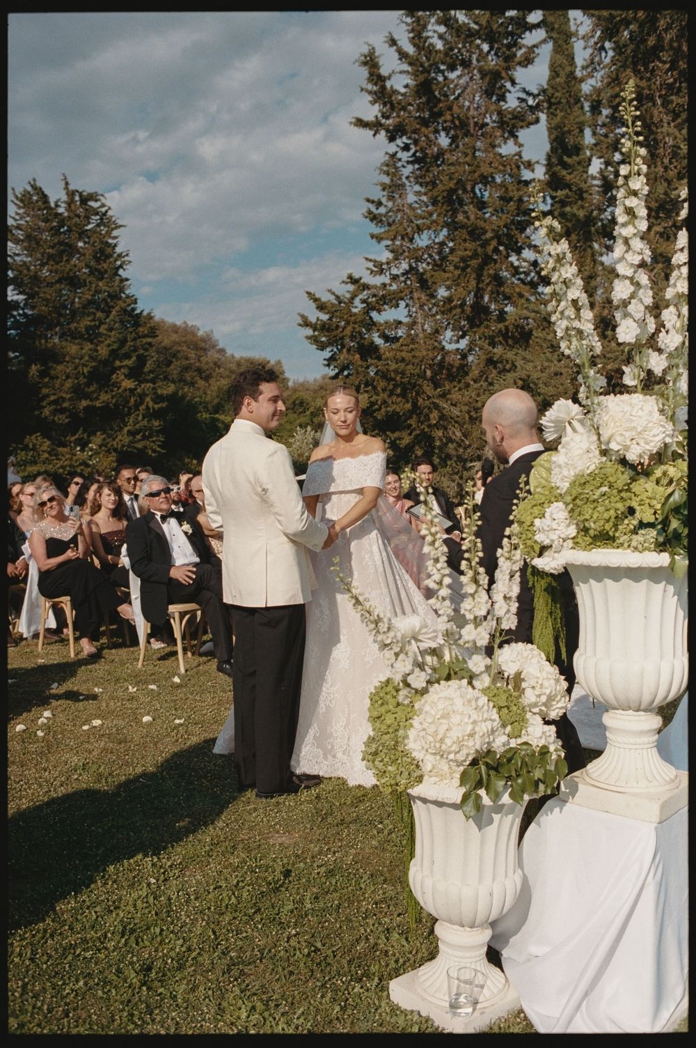 Outdoor wedding ceremony with couple exchanging vows at floral arch, white urn arrangements, guests seated on lawn.