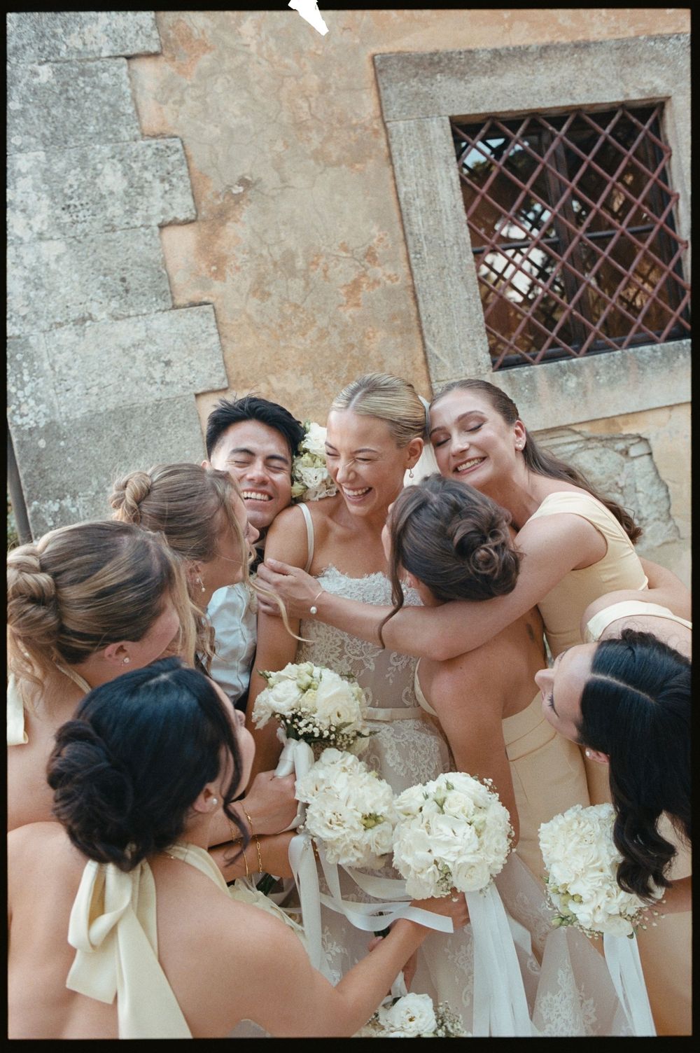Joyful bride surrounded by bridesmaids in champagne dresses sharing a group hug at stone venue.