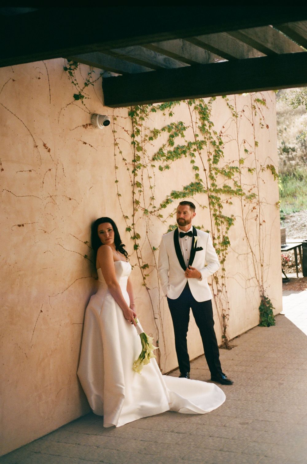 Bride in white gown and groom in white tuxedo pose against ivy-covered wall at elegant outdoor wedding venue.