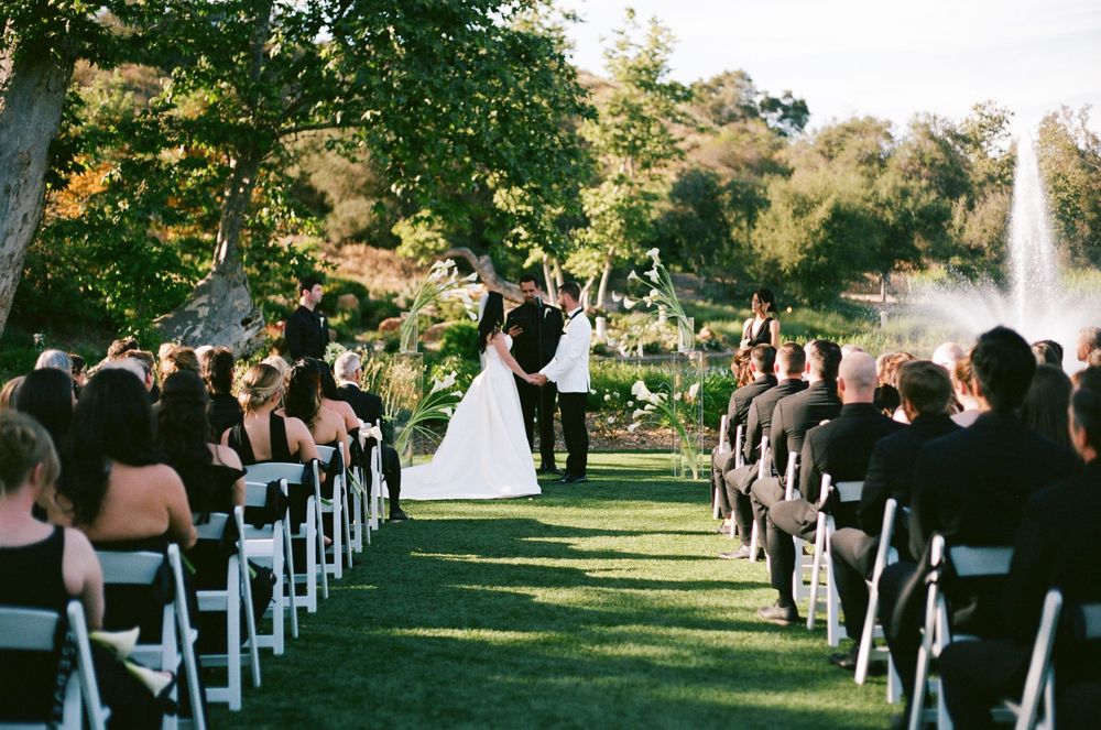 Outdoor wedding ceremony on lush green lawn with white chairs, fountain, and couple at the altar.