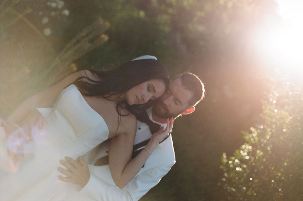 A bride and groom share a romantic embrace outdoors, bathed in warm golden sunlight filtering through lush green trees.