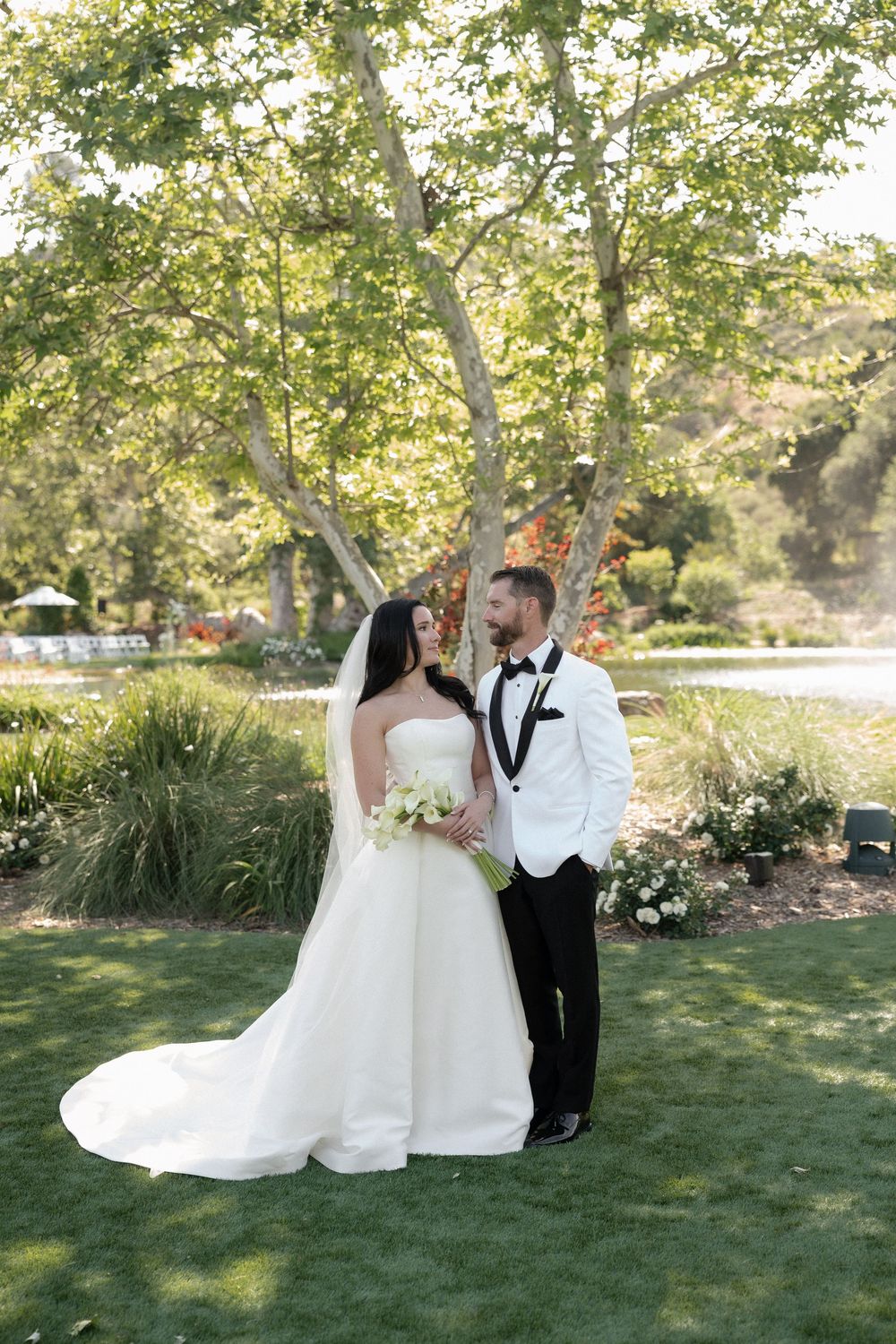Bride and groom share a romantic moment on green lawn surrounded by lush trees at outdoor wedding.