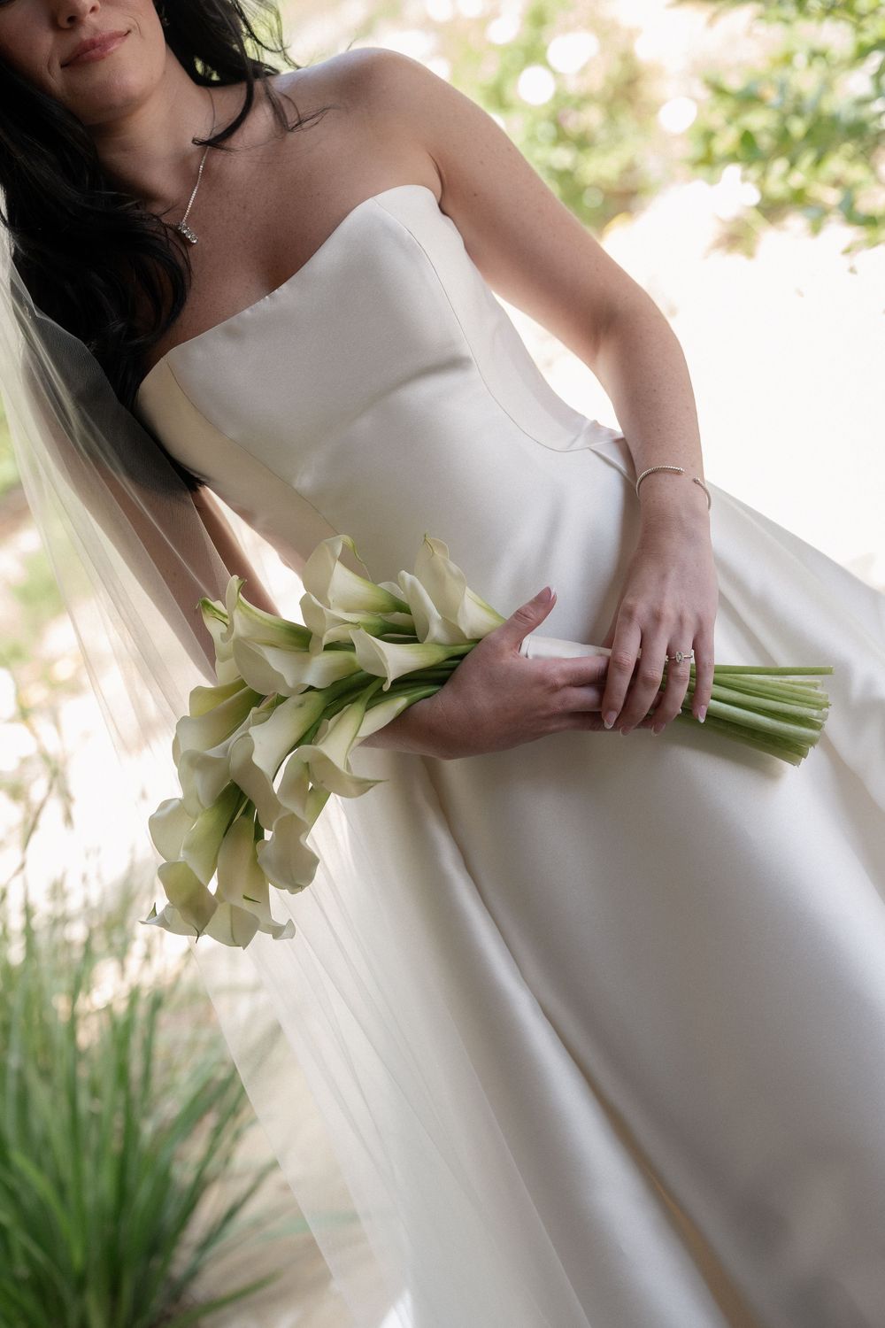Bride in a strapless white satin gown holding an elegant bouquet of white calla lilies with a flowing veil.