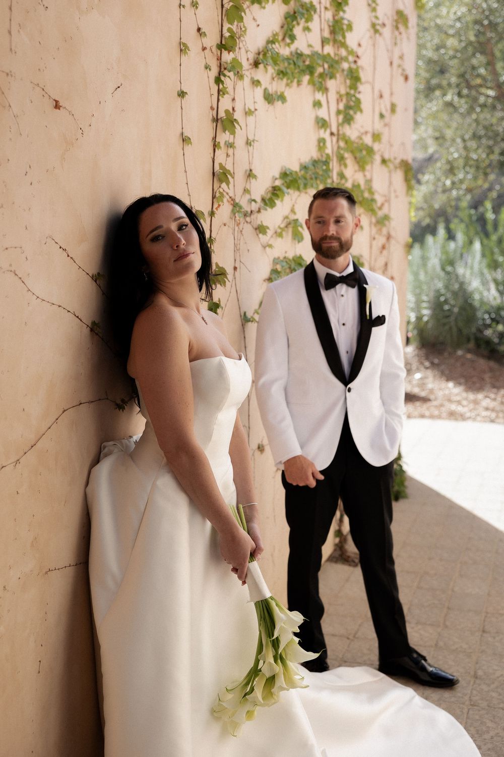 Bride leaning against a textured wall holding calla lilies while groom stands behind her.