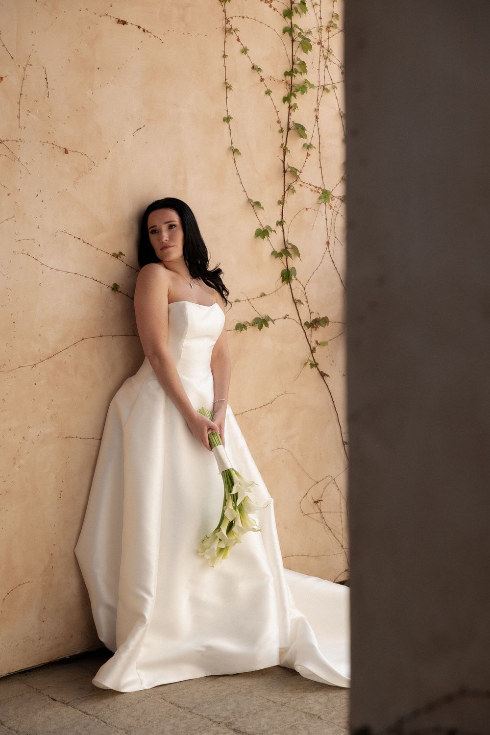Elegant bride in strapless white satin ballgown holding calla lilies, leaning against a textured wall with ivy.