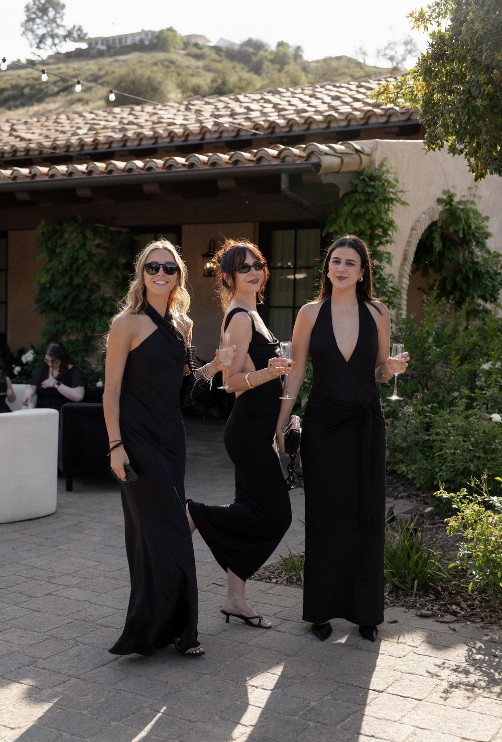 Three women in black formal gowns posing outdoors at a wedding reception venue.