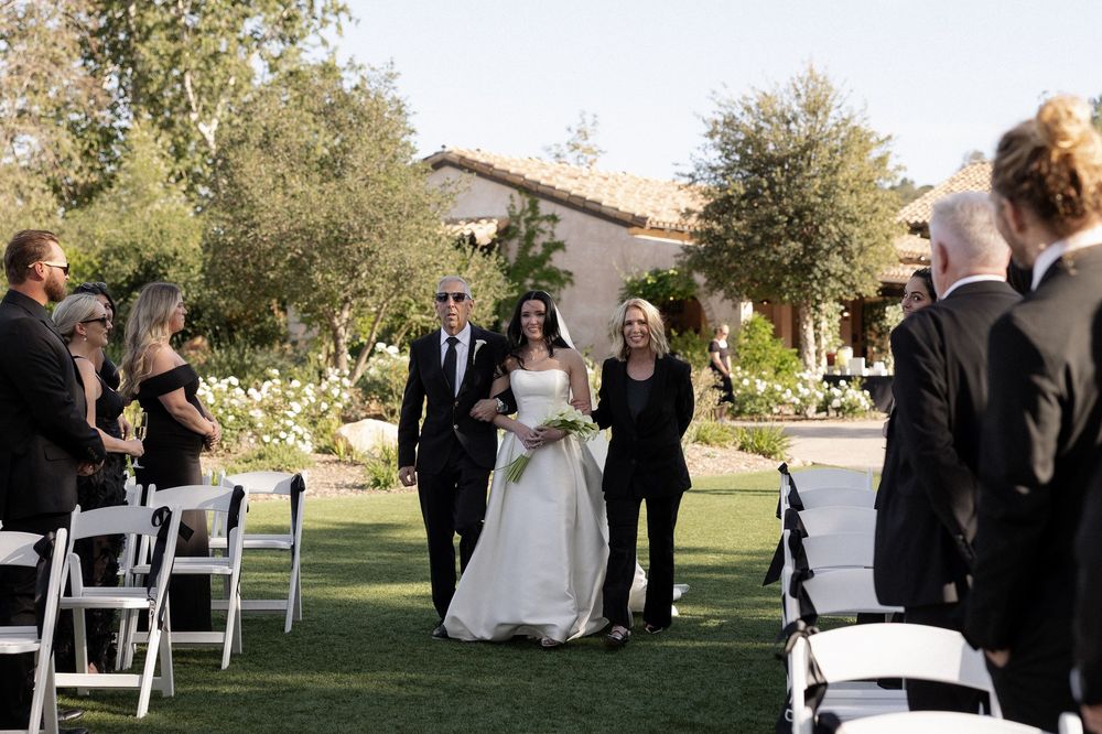 Bride walking down outdoor wedding aisle escorted by parents at a scenic garden venue.