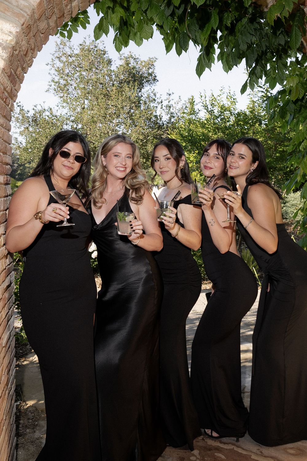 Five bridesmaids in elegant black satin gowns holding champagne glasses under a garden arch.