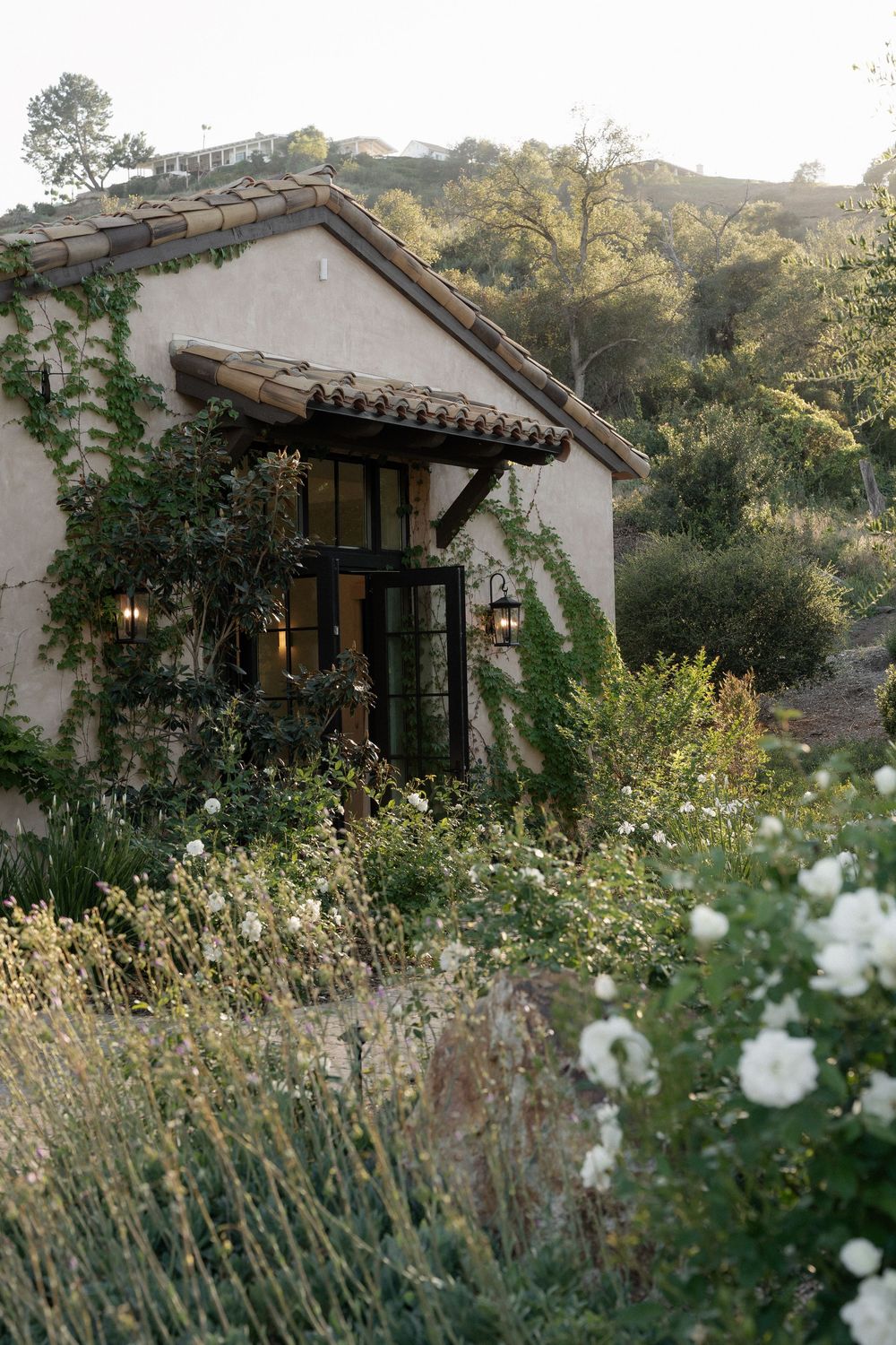 Ivy-covered Spanish-style cottage surrounded by wildflowers and lush gardens in a scenic hillside setting.