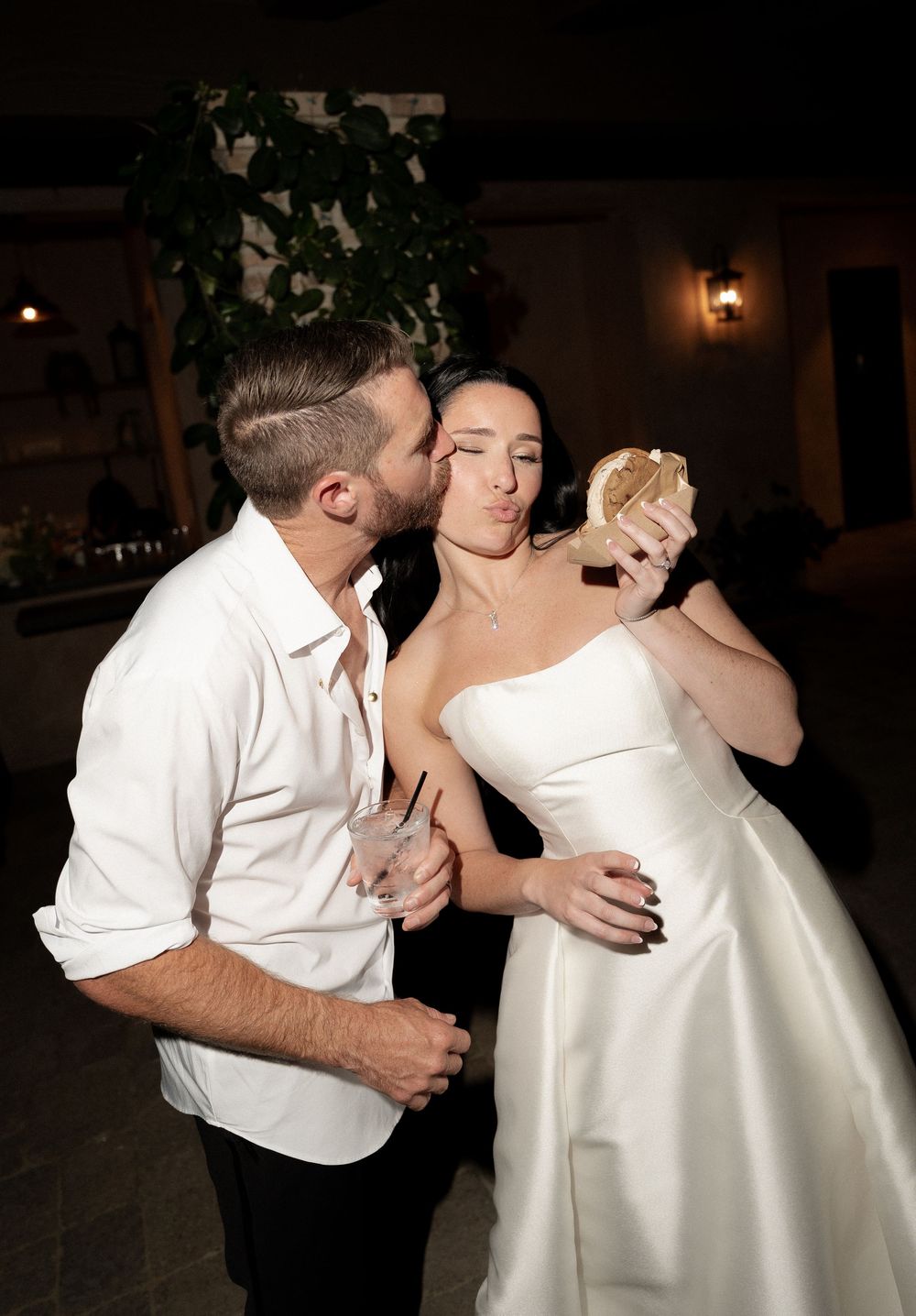 Bride in strapless white gown checks mirror while groom in white shirt kisses her cheek at evening reception.