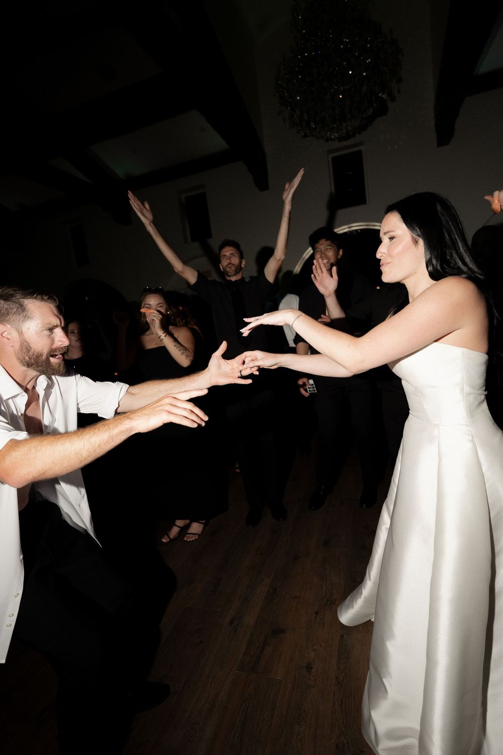 Bride in white gown reaches toward groom on wedding dance floor surrounded by celebrating guests.