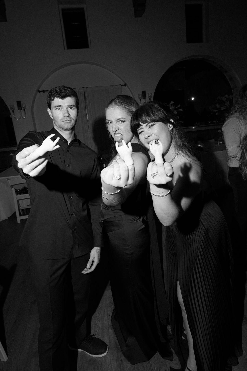 Three wedding guests in black attire pose playfully showing rings during a black and white reception photo.