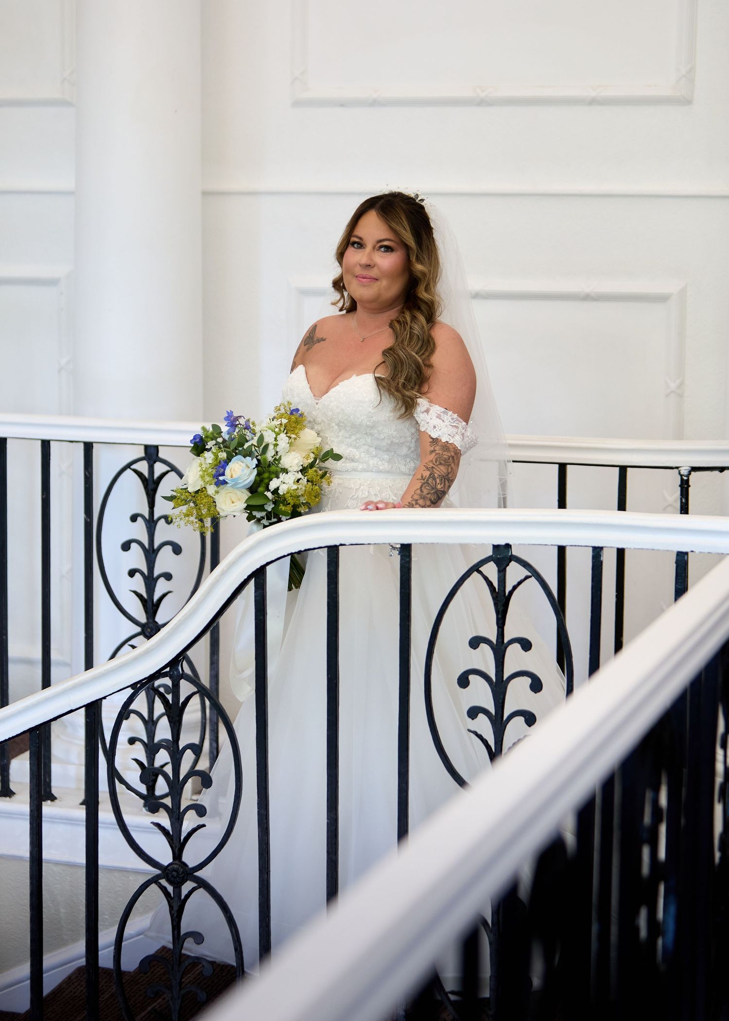 Bride in white off-shoulder gown holding blue bouquet stands on elegant staircase with ornate iron railings.