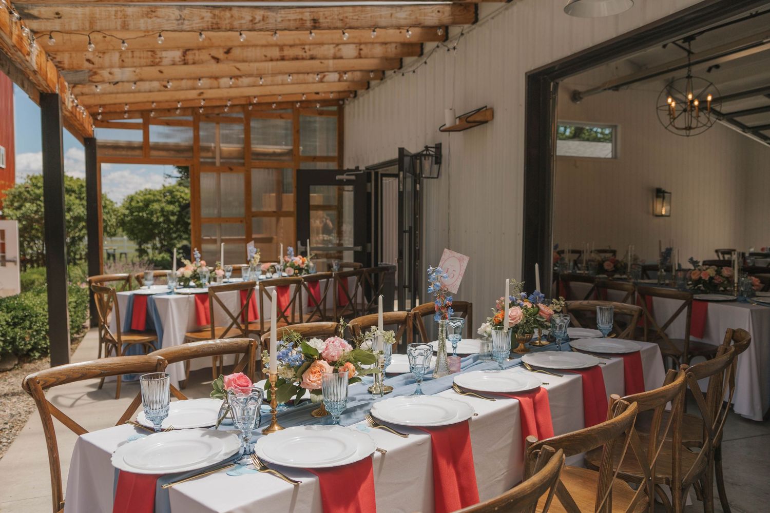 Elegant outdoor wedding reception with long tables, red napkins, floral centerpieces, and rustic wooden chairs under a pergola.