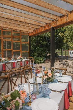 Elegant outdoor wedding reception setup with wooden pergola, floral centerpieces, blue table runner, and white place settings.