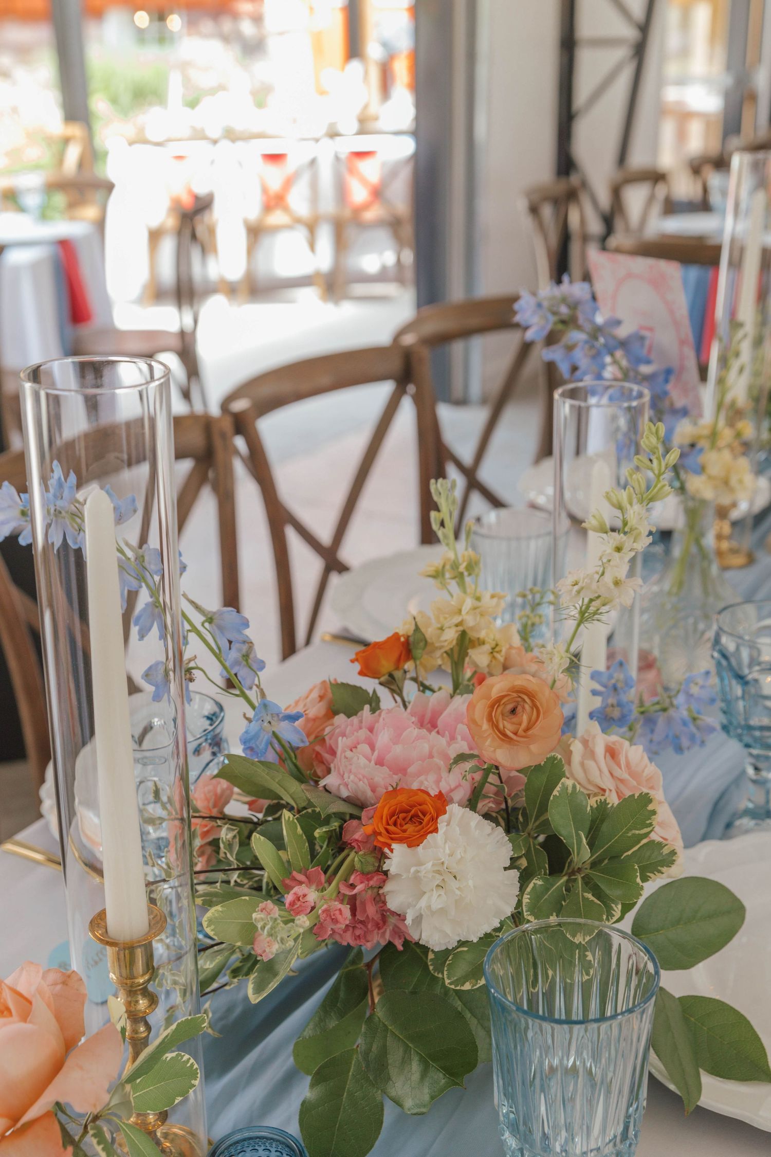 Elegant wedding table centerpiece with colorful blooms, blue glassware, and candles in a bright reception venue.