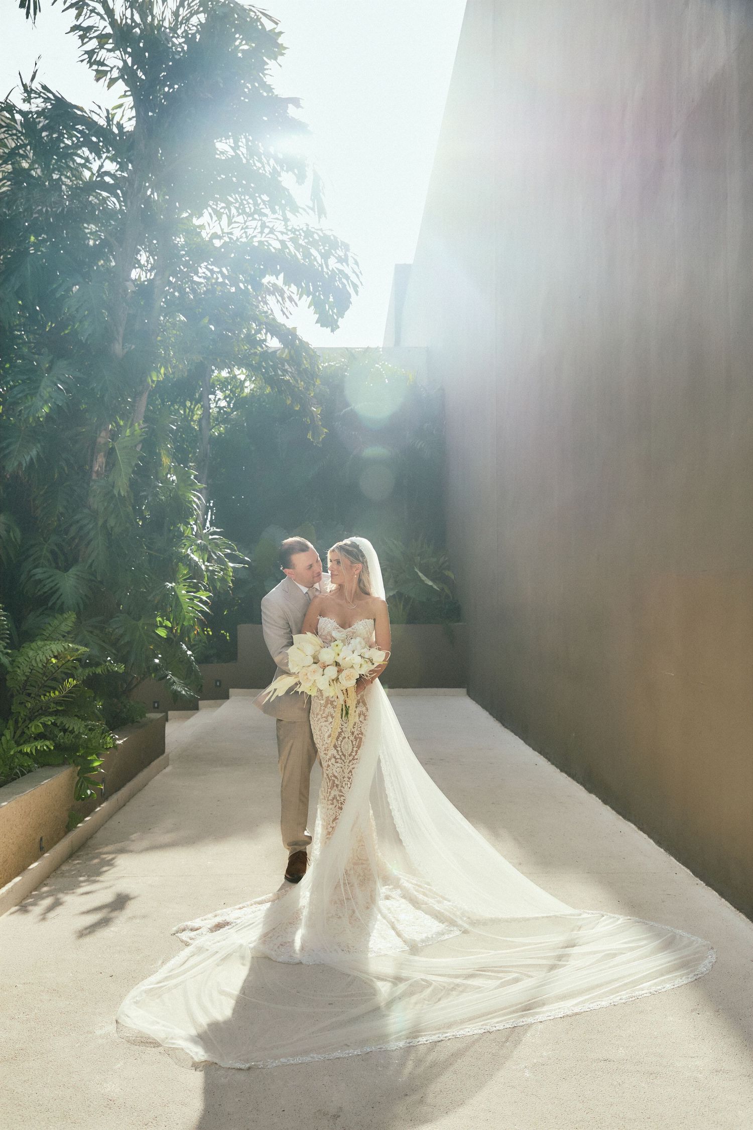 Bride and groom walk in sunlit alley, her long veil trailing behind, bouquet in hand.