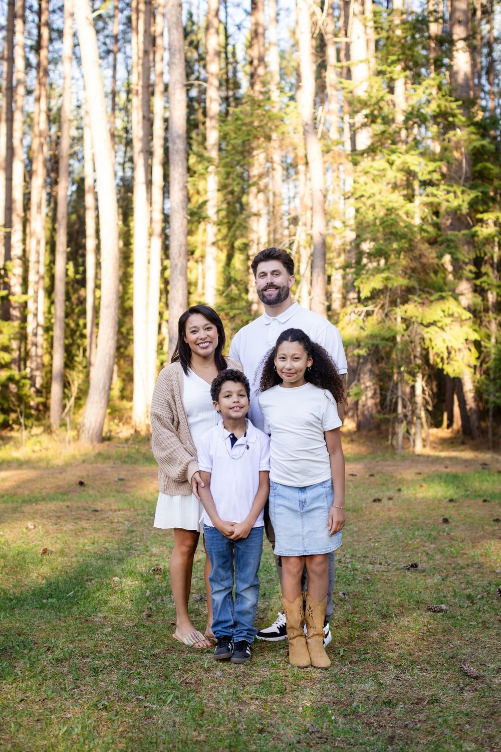 Happy family of four posing together in a sunlit pine forest, smiling for a portrait outdoors.