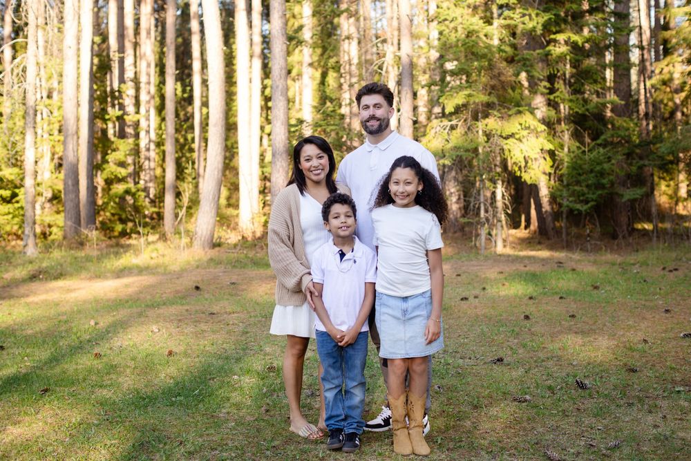 Family of four posing outdoors in a sunlit forest clearing, parents and two children smiling in coordinated white outfits.