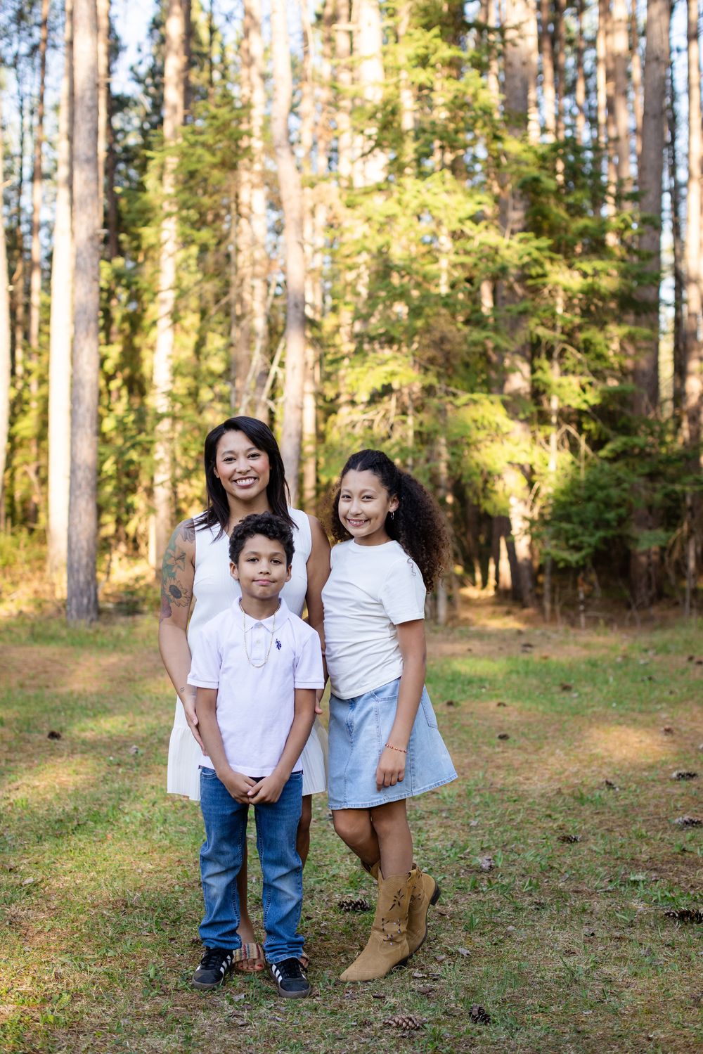 A smiling woman and two children pose together in a sunlit forest clearing, all wearing white tops with casual bottoms.
