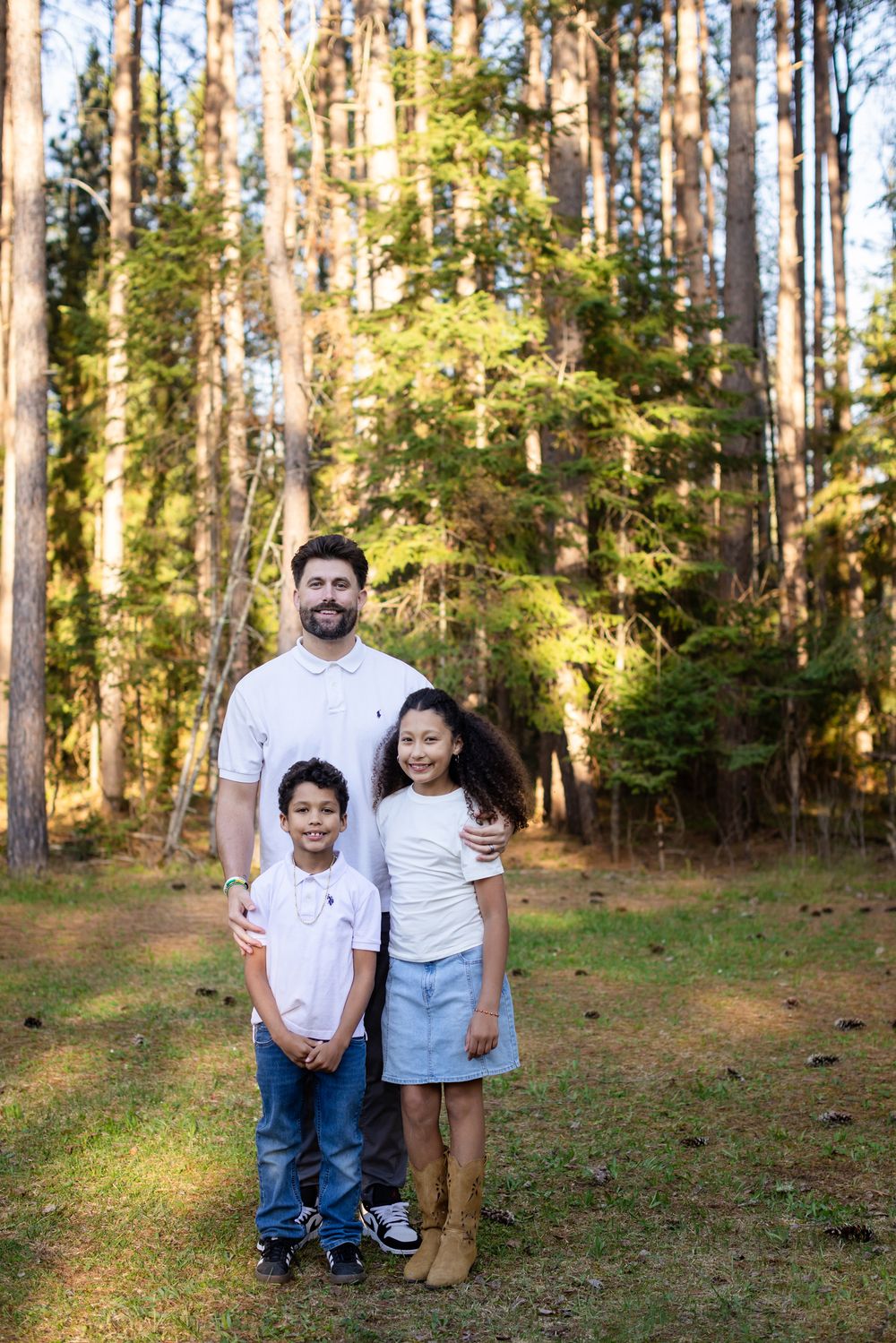 A father poses with his two children in a sunlit forest clearing, all wearing white tops and casual attire.