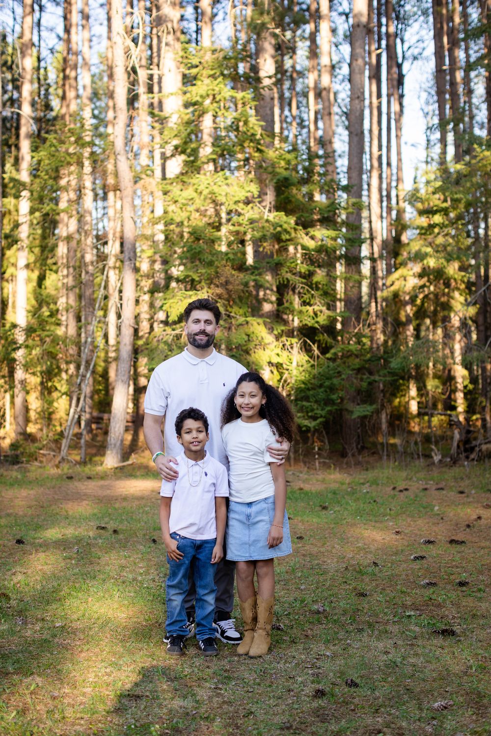 A man and two children smile together in a sunny woodland setting, dressed in coordinating white shirts and jeans.