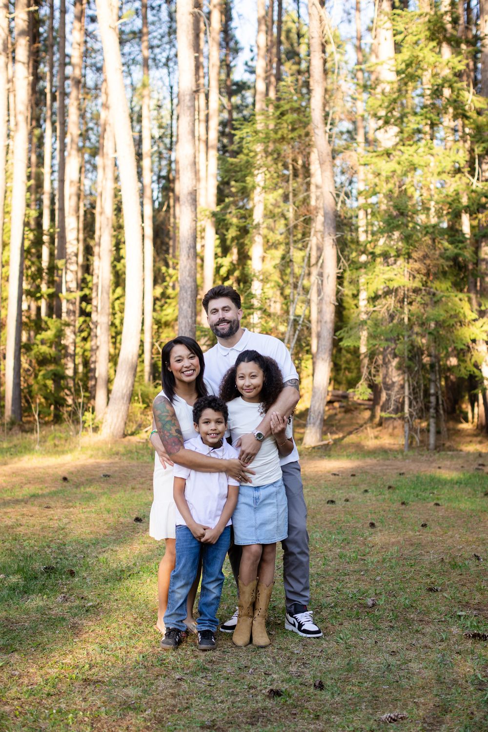 Happy interracial family of four posing together in a sunny pine forest, smiling for a portrait outdoors.