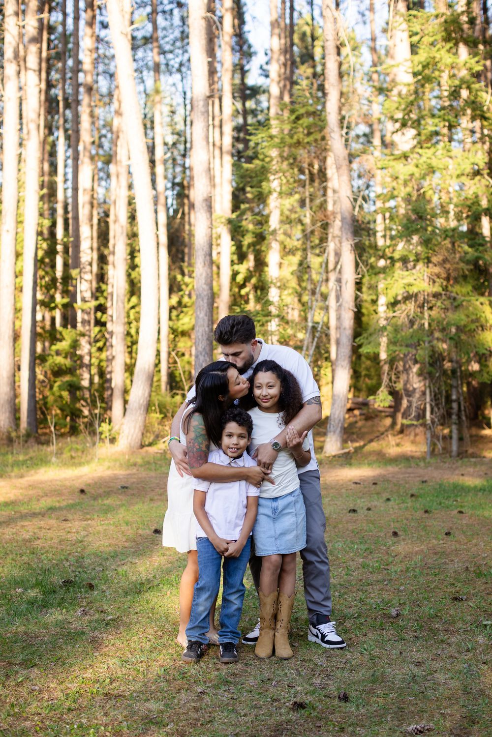 A happy family of four shares hugs and kisses in a sunlit forest clearing, with tall pine trees in the background.