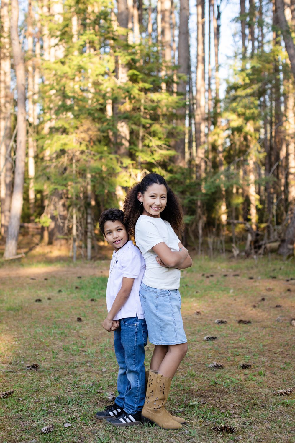 Two children in white shirts stand back-to-back smiling in a sunlit forest clearing with pine trees.