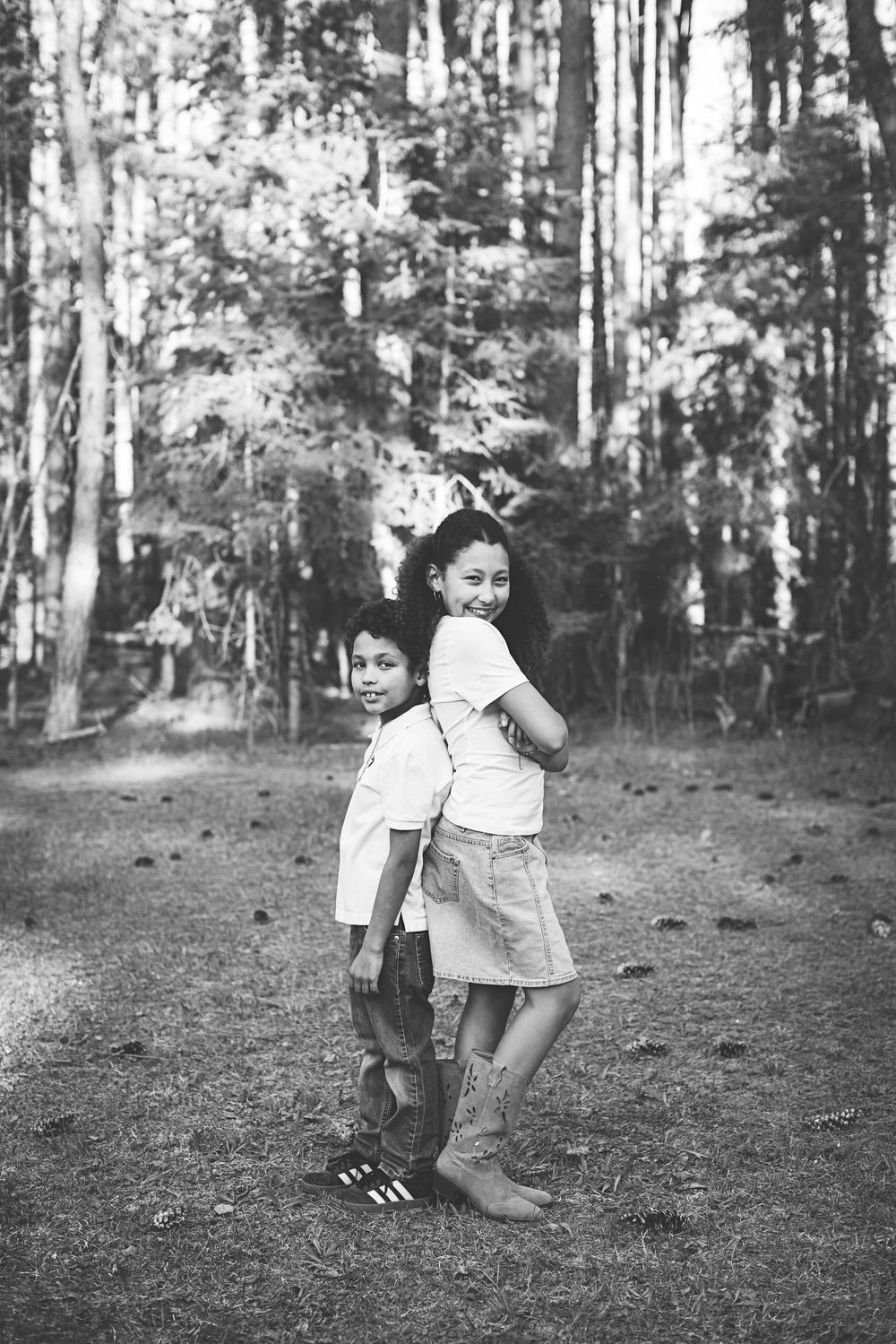 Two children stand back-to-back smiling in a forest, black and white photo.