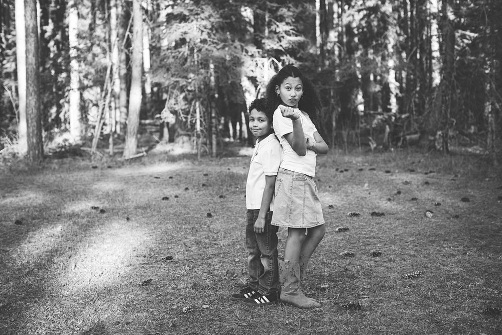Black and white photo of two children embracing in a forest setting, standing on a dirt path surrounded by tall trees.