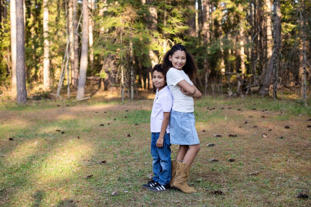Two children pose back-to-back smiling in a sunny forest clearing, girl in denim skirt hugging boy in white shirt.