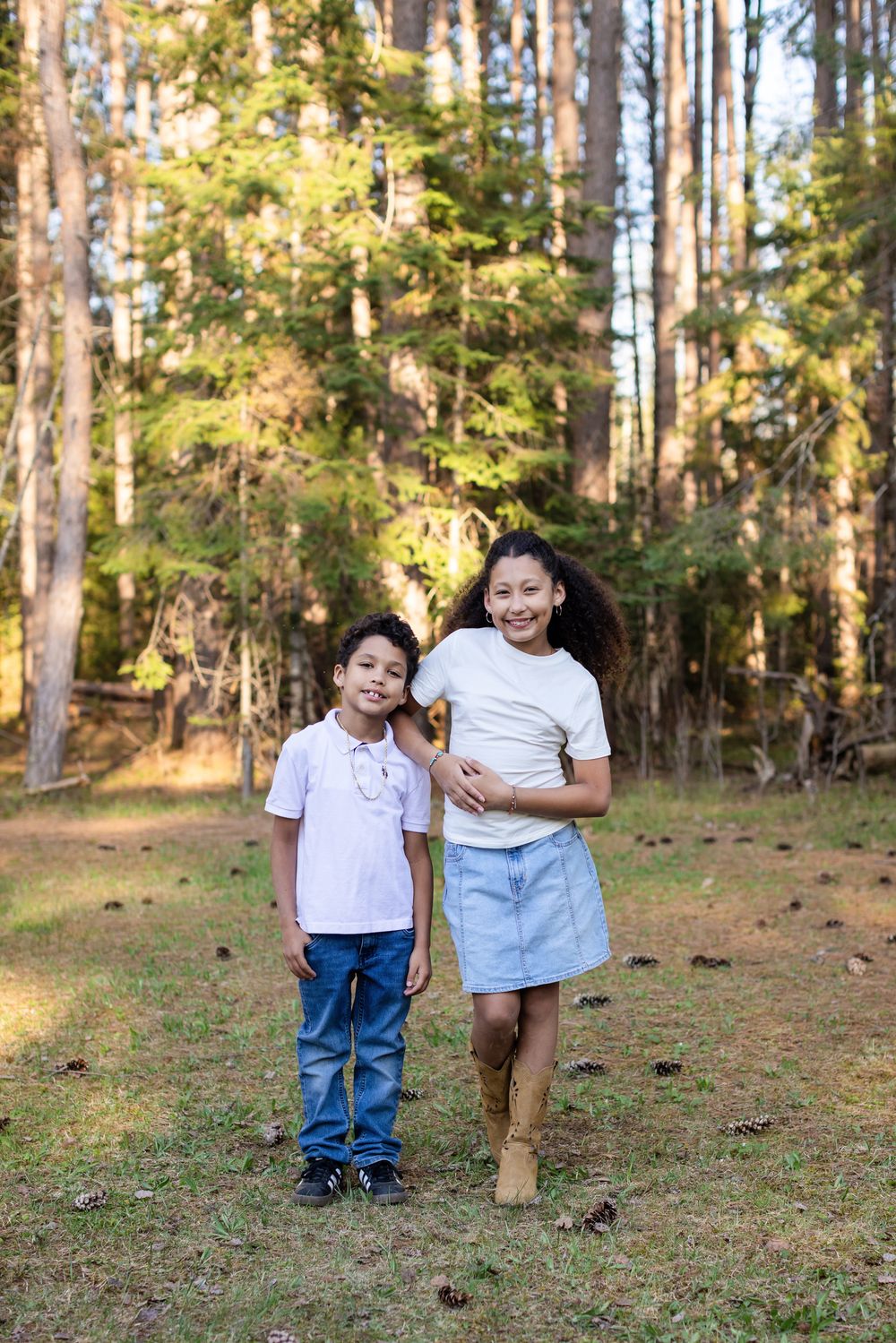 Two children in white shirts pose smiling in a sunlit forest, girl in denim skirt and boots with younger boy.