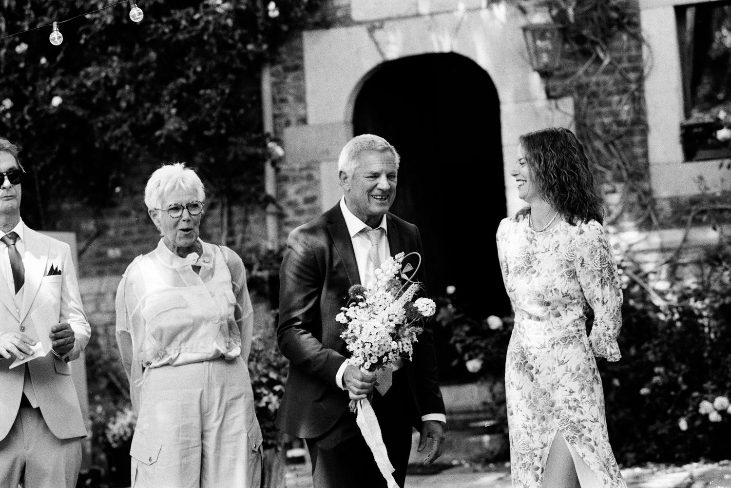 Black and white photo of a bride holding a bouquet walking with a man in a suit at a wedding ceremony.