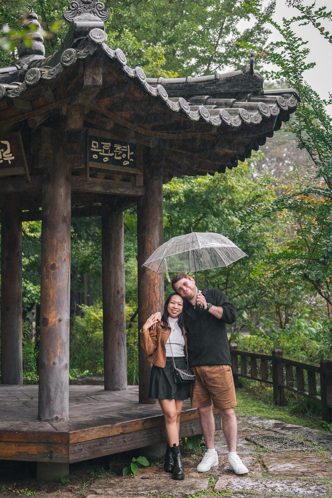 Couple sharing a clear umbrella by a traditional Korean pavilion surrounded by lush greenery on a rainy day.