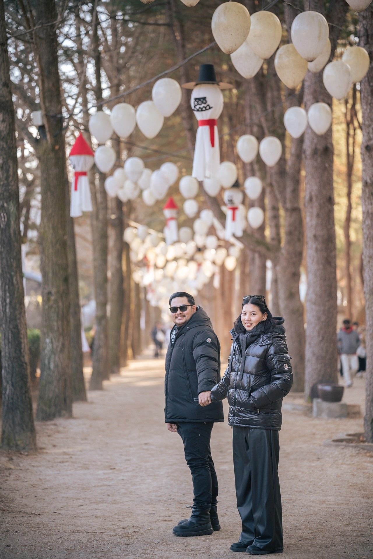 Couple holding hands on a tree-lined path decorated with white balloons and snowman ornaments in winter.