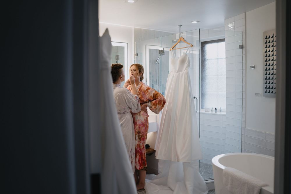 Two women in robes admire a white wedding dress hanging in an elegant bathroom before the ceremony.