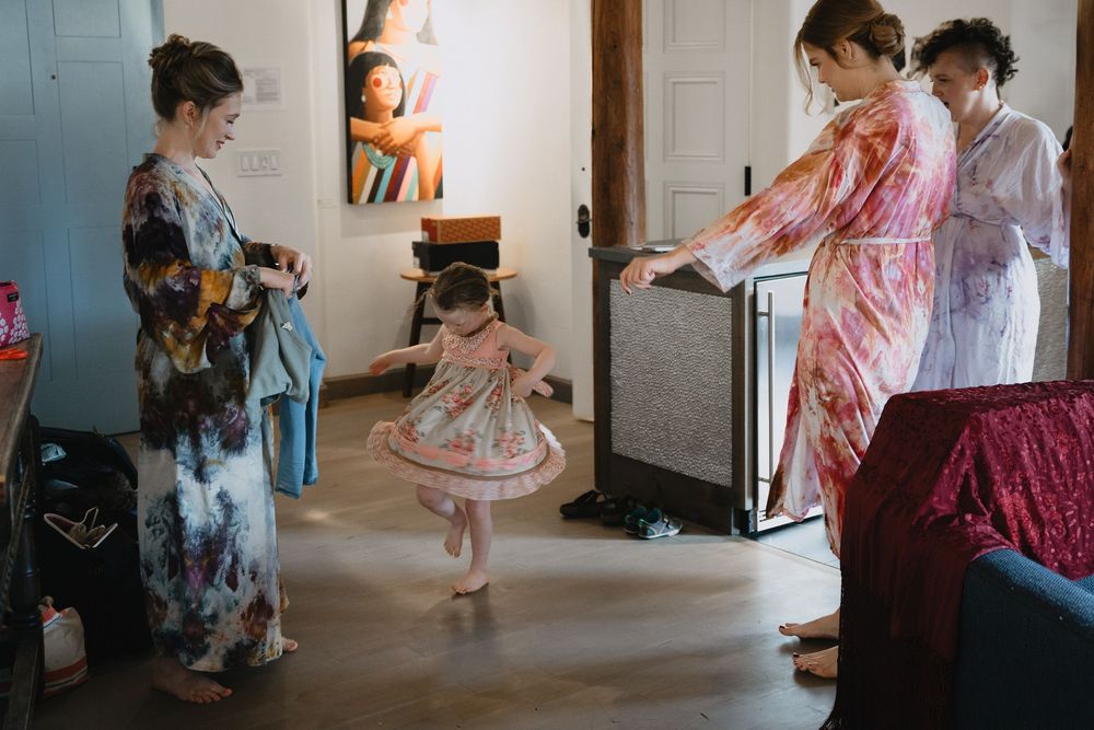 A little girl in a floral dress twirls while women in robes prepare for a wedding getting-ready celebration indoors.