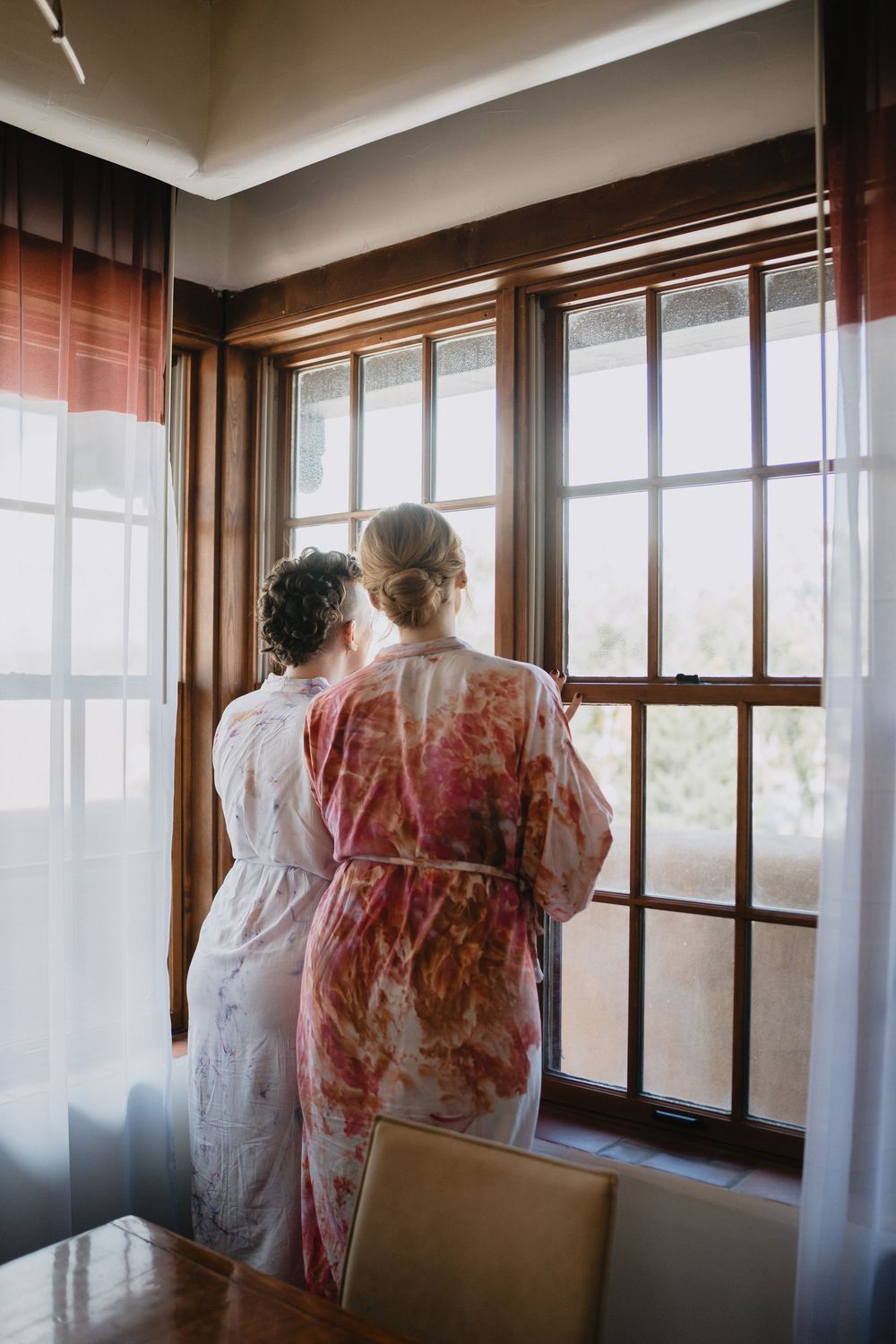 Two women in floral robes stand together looking out a wooden-framed window in a softly lit room.