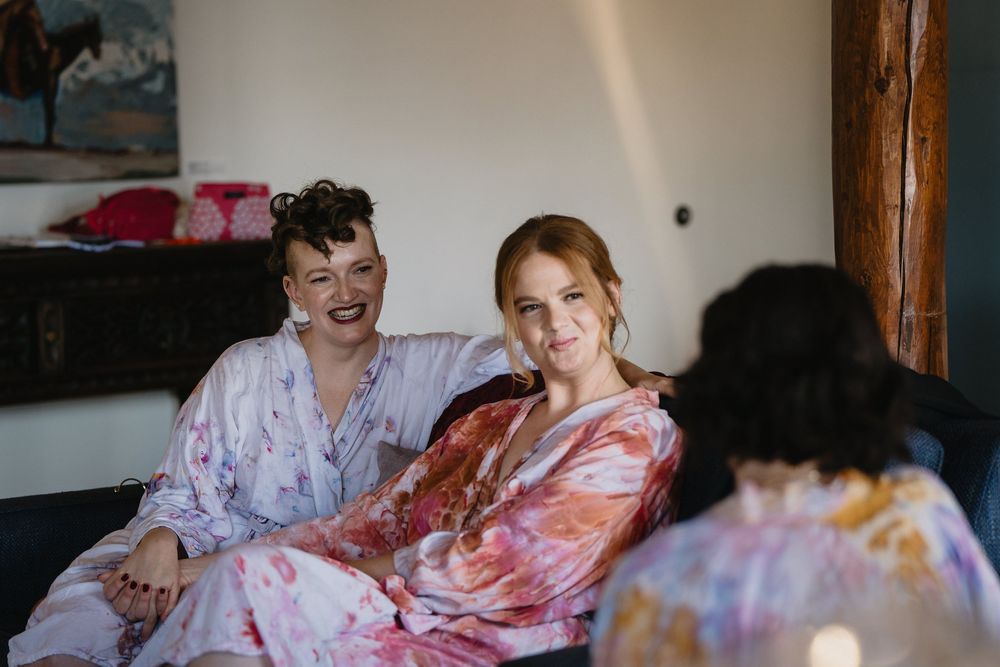 Two women in colorful floral robes smile and laugh together while sitting on a couch during bridal preparations.