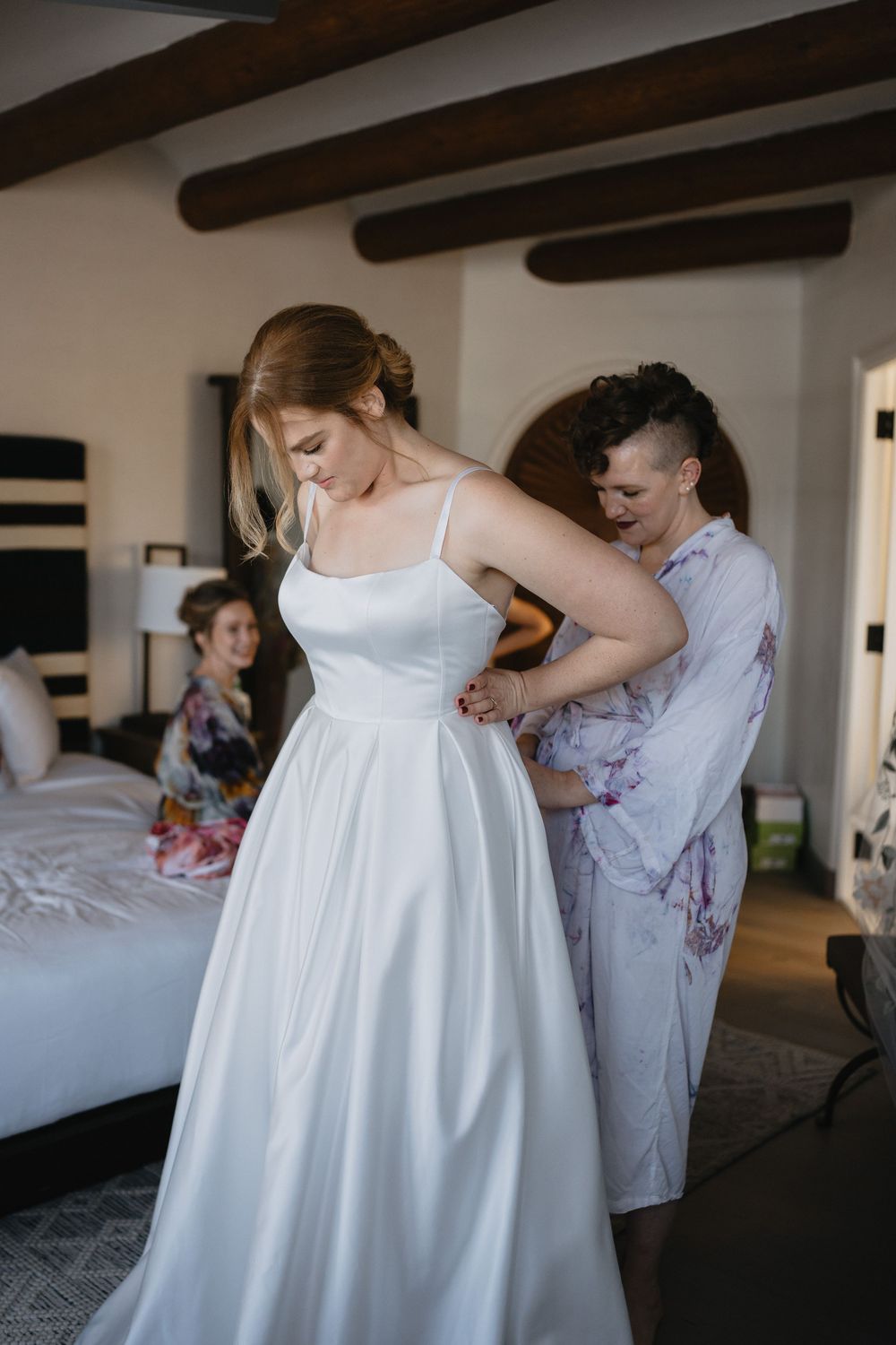 Bride getting help with her wedding dress from a bridesmaid in a floral robe in an elegant room.